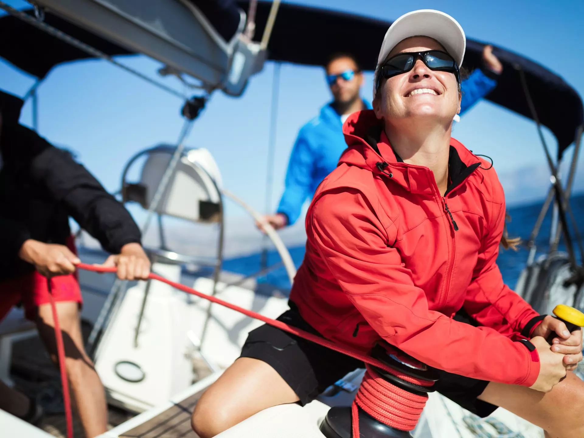 Eine Frau in roter Segeljacke und weißer Kappe bedient eine Winsch an Bord einer Segelyacht.