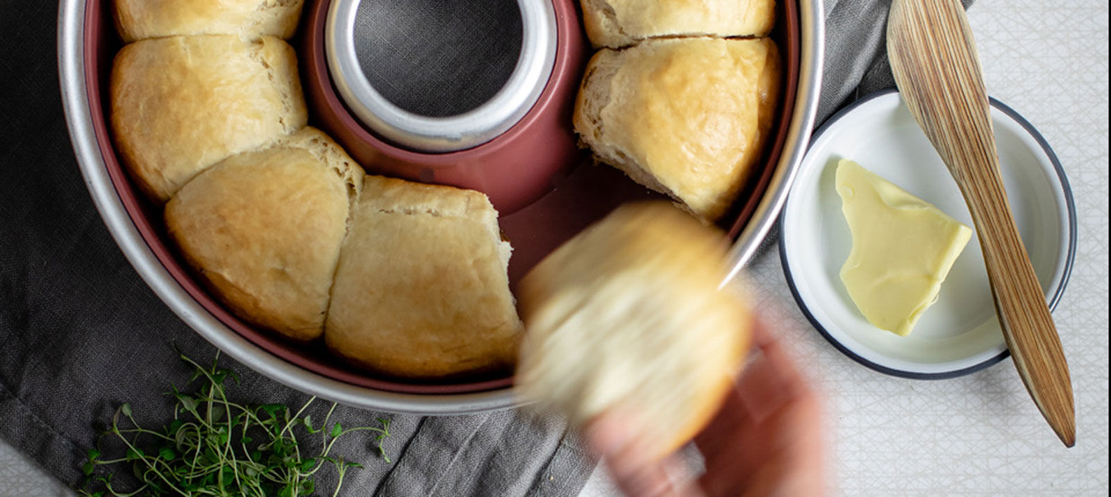 Eine Hand nimmt ein frisch gebackenes Brötchen aus einer roten Omnia Backform, daneben Butter und Kräuter.