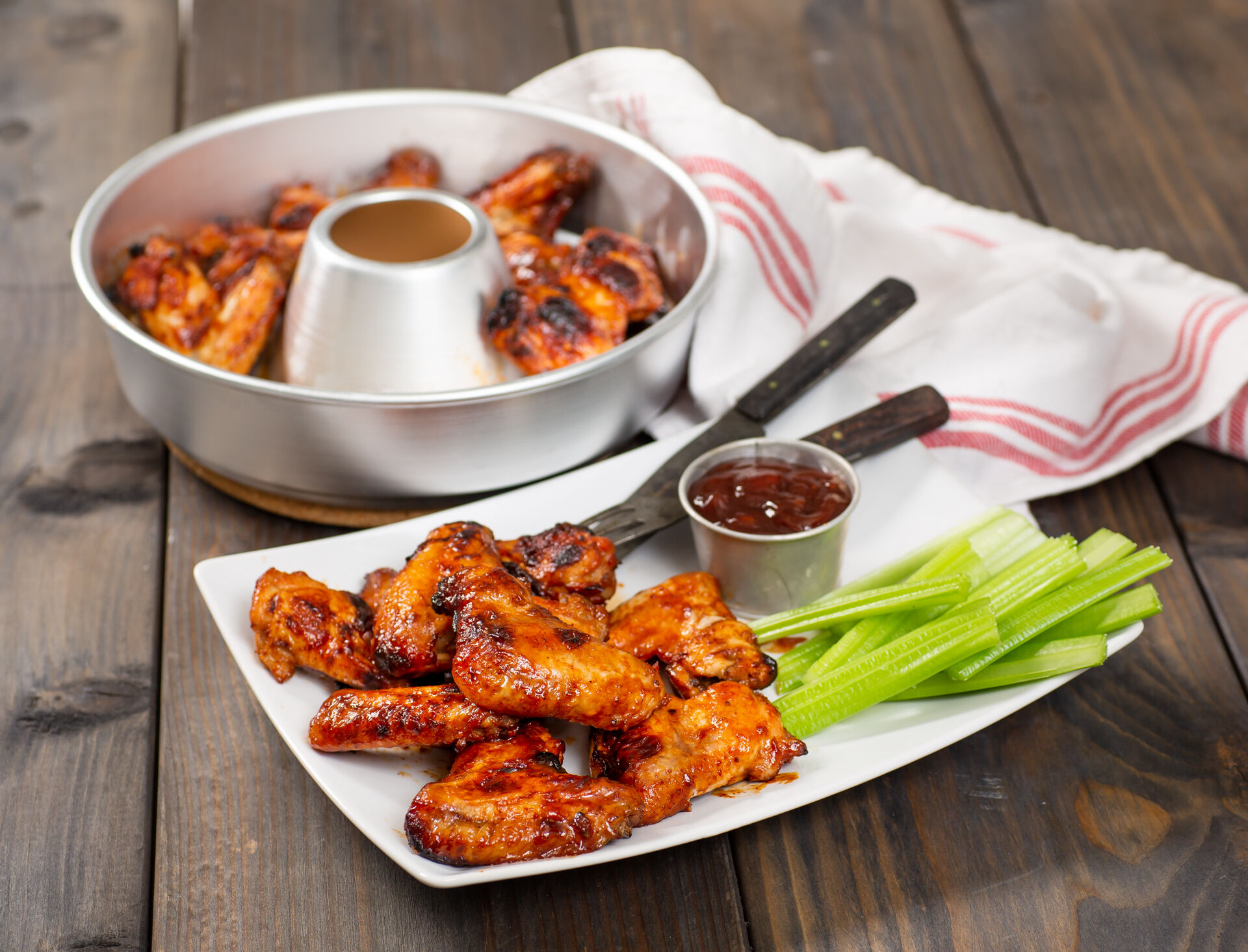 Gegrillte Chicken Wings auf einem Teller mit Sellerie und Dip sowie eine runde Backform im Hintergrund